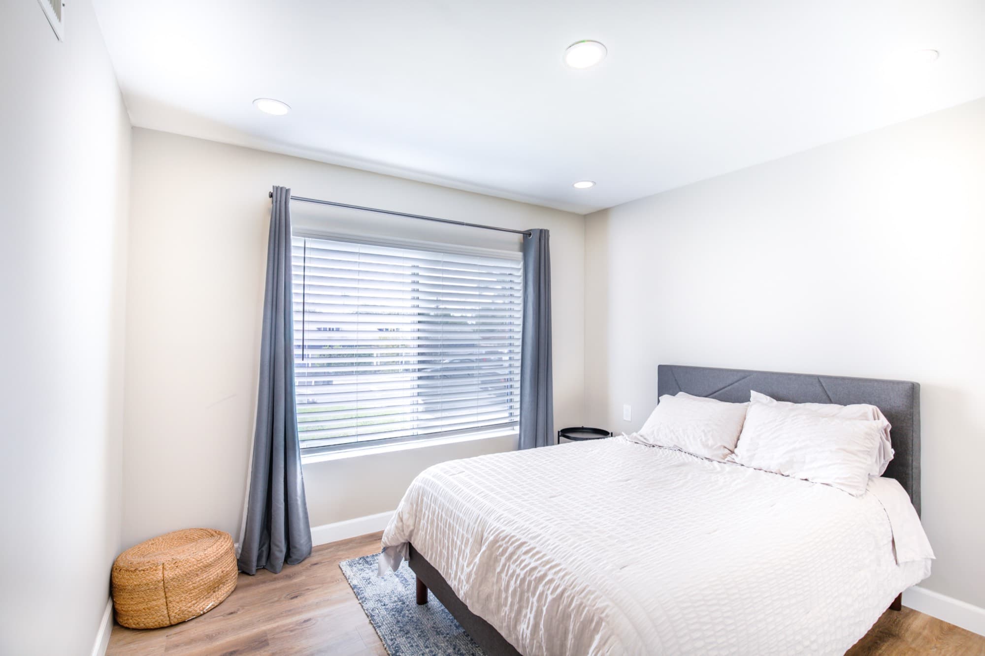 Guest bedroom with queen bed, grey curtains, woven pouf, and hardwood floors