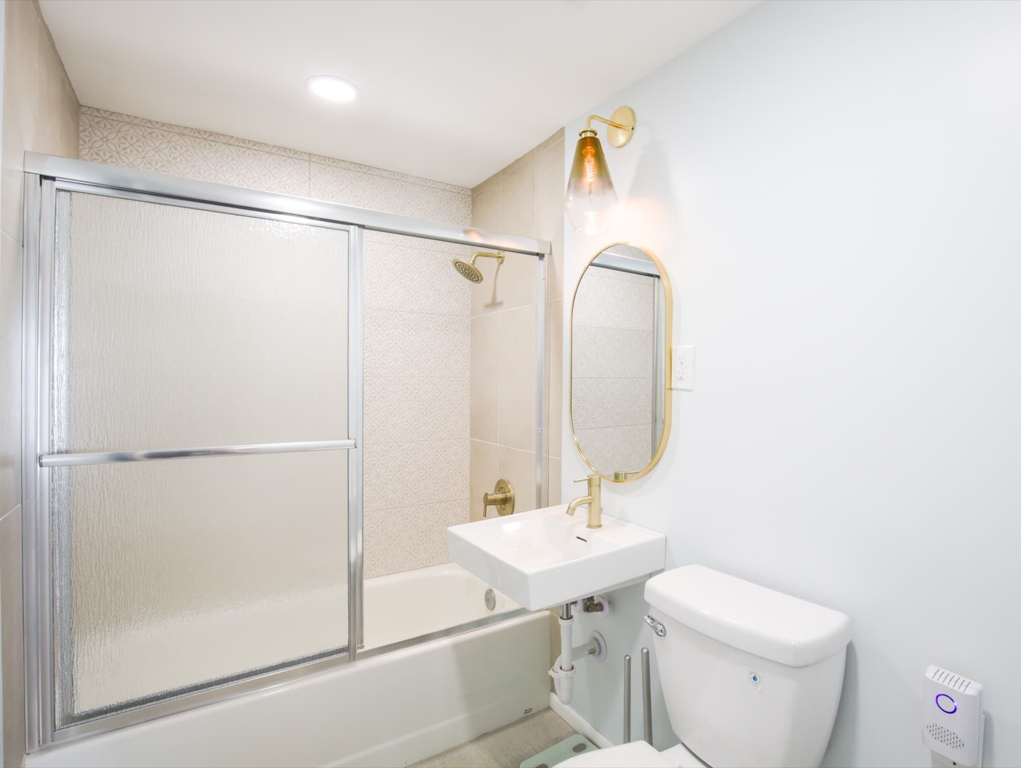 En-suite bathroom with gold fixtures, oval mirror, patterned tile shower, and pedestal sink